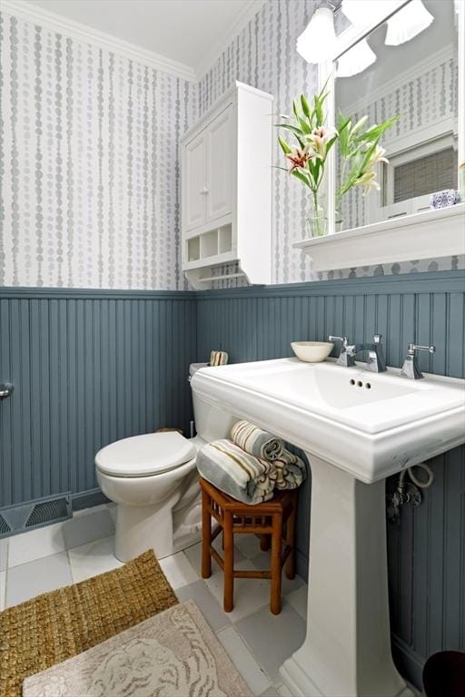 half bathroom with wallpapered walls, crown molding, toilet, tile patterned floors, and wainscoting