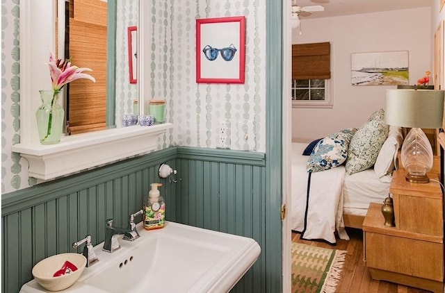 bathroom featuring wainscoting, a sink, wood finished floors, a ceiling fan, and wallpapered walls