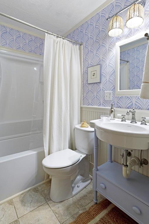 bathroom with wallpapered walls, toilet, wainscoting, shower / bath combination with curtain, and tile patterned floors