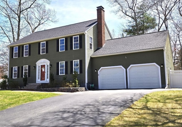 colonial-style house with a chimney, aphalt driveway, a shingled roof, a garage, and a front yard