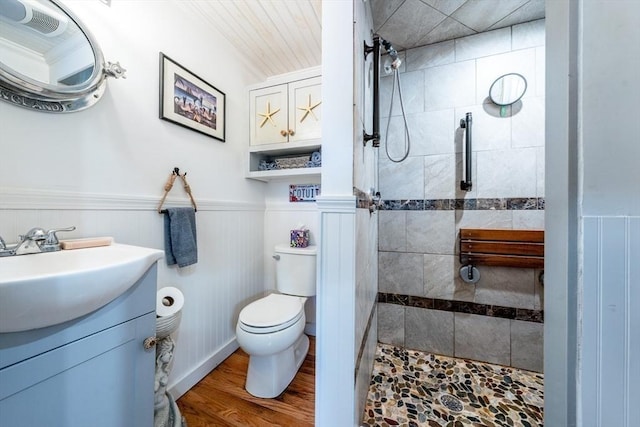bathroom with toilet, wood finished floors, vanity, wainscoting, and tiled shower
