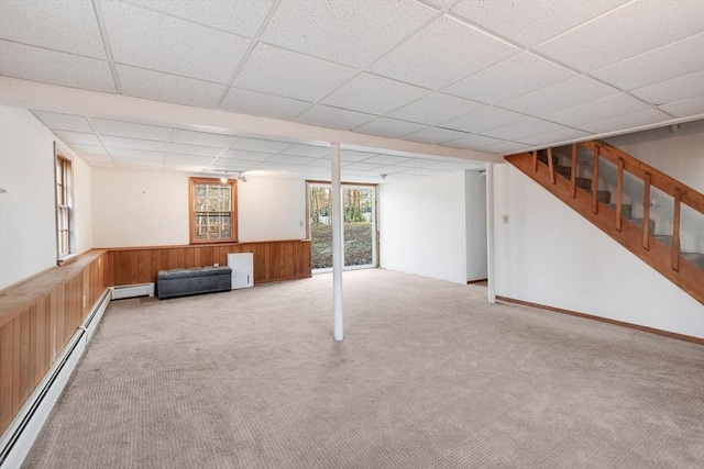 basement featuring a wainscoted wall, wooden walls, a paneled ceiling, baseboard heating, and stairs