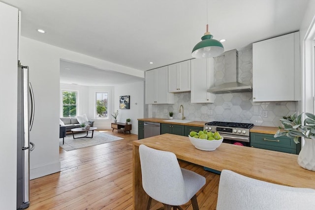 kitchen with wall chimney range hood, stainless steel appliances, wood counters, a sink, and white cabinets
