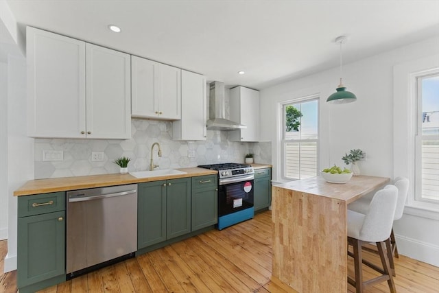 kitchen with stainless steel appliances, wall chimney range hood, a sink, green cabinetry, and butcher block countertops