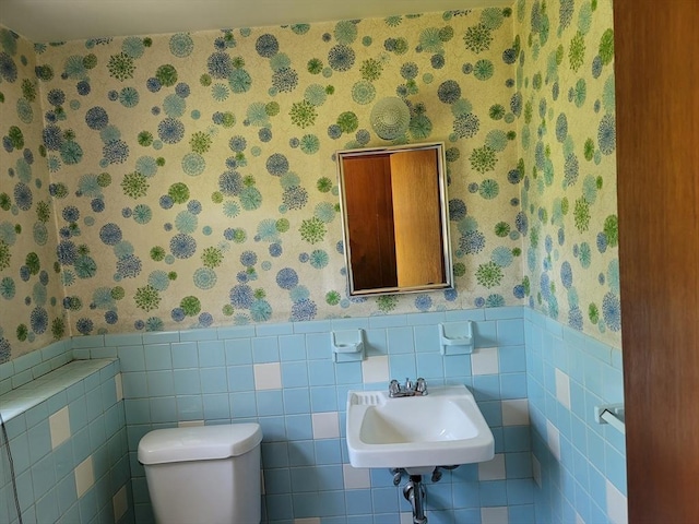 half bathroom with wallpapered walls, a sink, toilet, wainscoting, and tile walls