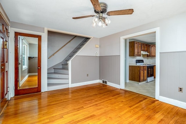 unfurnished living room with light wood-style floors, a ceiling fan, stairs, a wainscoted wall, and baseboards