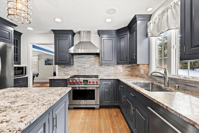 kitchen with pendant lighting, stainless steel appliances, a sink, wall chimney range hood, and light wood-type flooring