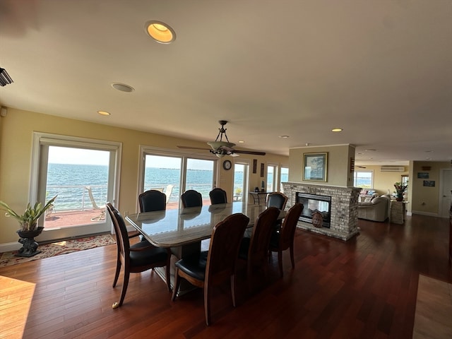 dining room with a water view, ceiling fan, dark wood-type flooring, and a stone fireplace