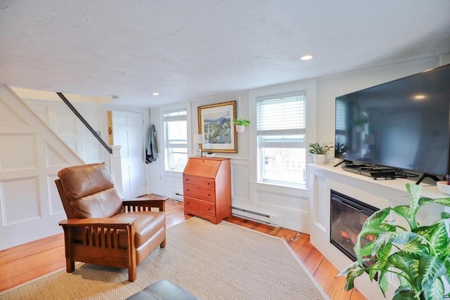 sitting room with stairs, a decorative wall, a baseboard radiator, light wood-style flooring, and a glass covered fireplace