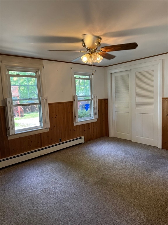 unfurnished bedroom featuring baseboard heating, wooden walls, carpet floors, and ceiling fan