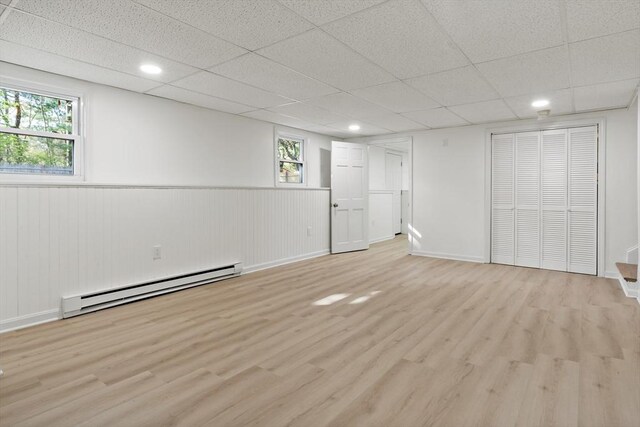 basement featuring a paneled ceiling, a baseboard radiator, wainscoting, and light wood-style floors