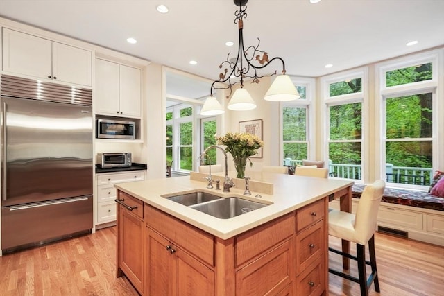 kitchen featuring built in appliances, a kitchen island with sink, a sink, white cabinetry, and decorative light fixtures