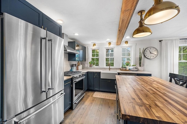 kitchen with high end appliances, a sink, wood counters, wall chimney exhaust hood, and blue cabinetry