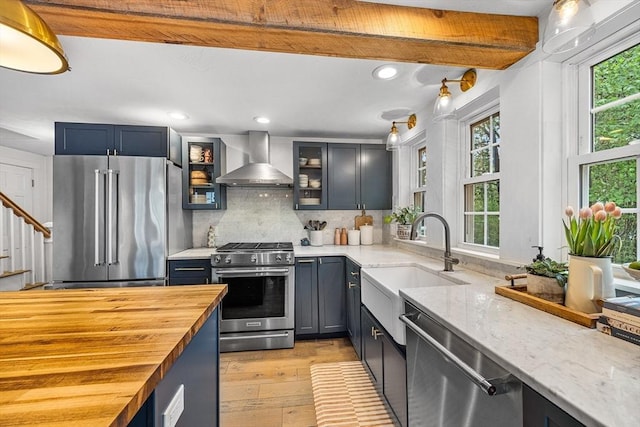 kitchen featuring appliances with stainless steel finishes, wall chimney range hood, butcher block counters, a sink, and light wood finished floors