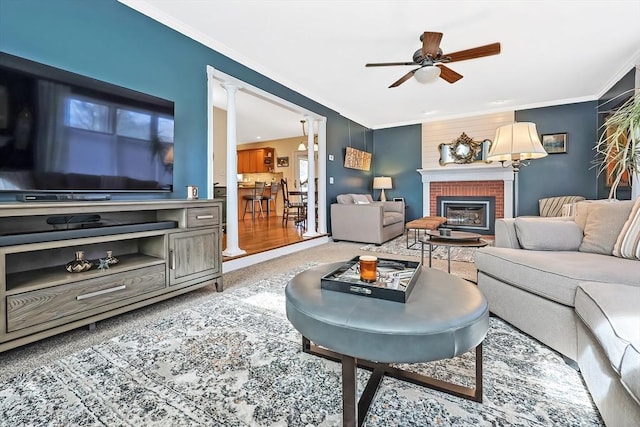 carpeted living room featuring a brick fireplace, crown molding, ceiling fan, and decorative columns