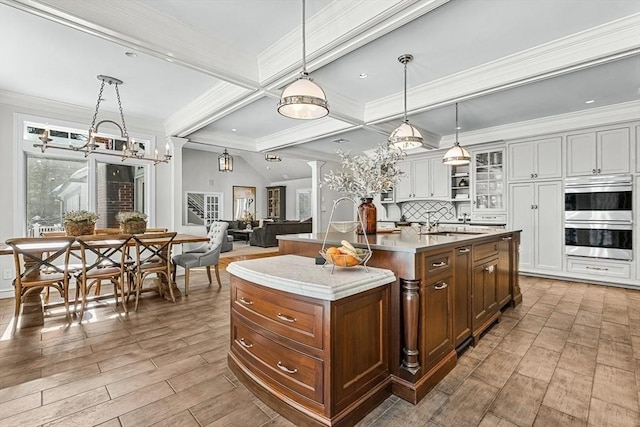 kitchen with beam ceiling, a center island with sink, double oven, light wood-style floors, and glass insert cabinets