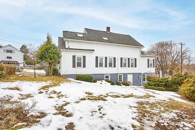 snow covered property with a shingled roof, a chimney, and stucco siding