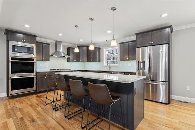 kitchen with wall chimney exhaust hood, hanging light fixtures, appliances with stainless steel finishes, a kitchen breakfast bar, and a kitchen island