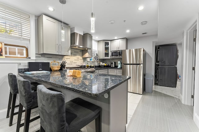 kitchen featuring tasteful backsplash, stainless steel appliances, wall chimney range hood, pendant lighting, and dark stone countertops