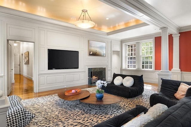 living room featuring light wood-style flooring, a decorative wall, a fireplace with flush hearth, ornate columns, and crown molding