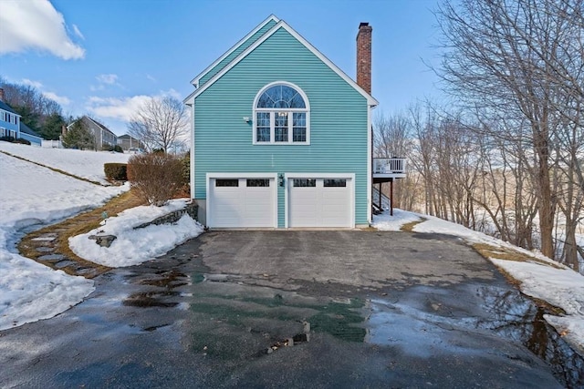 snow covered property featuring driveway, an attached garage, and a chimney