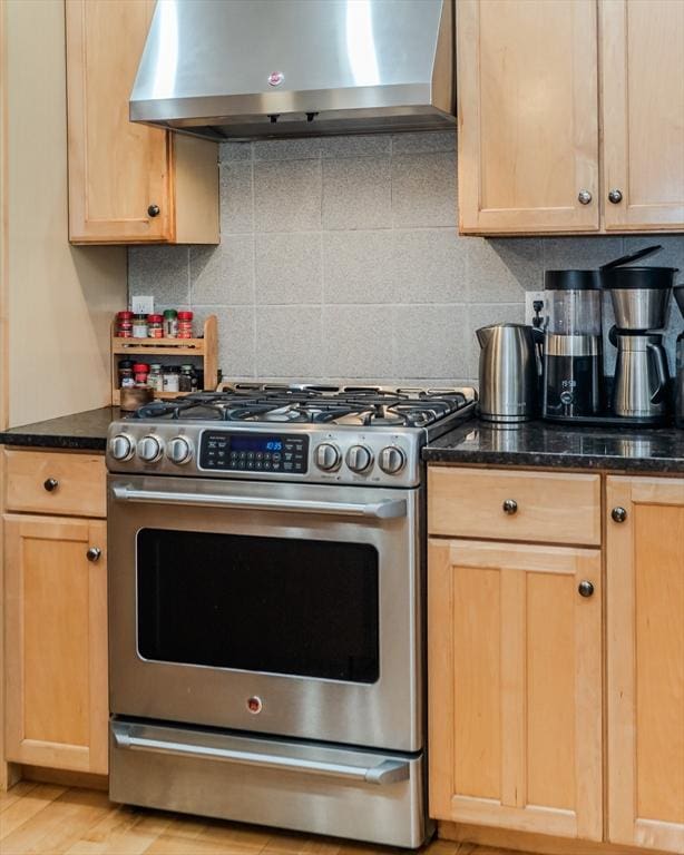 kitchen featuring backsplash, light brown cabinetry, stainless steel gas stove, dark stone countertops, and wall chimney exhaust hood