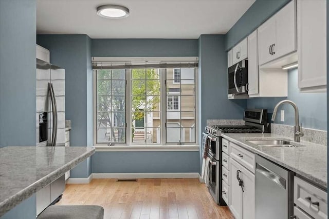 kitchen with appliances with stainless steel finishes, a sink, white cabinets, light wood-type flooring, and light stone counters