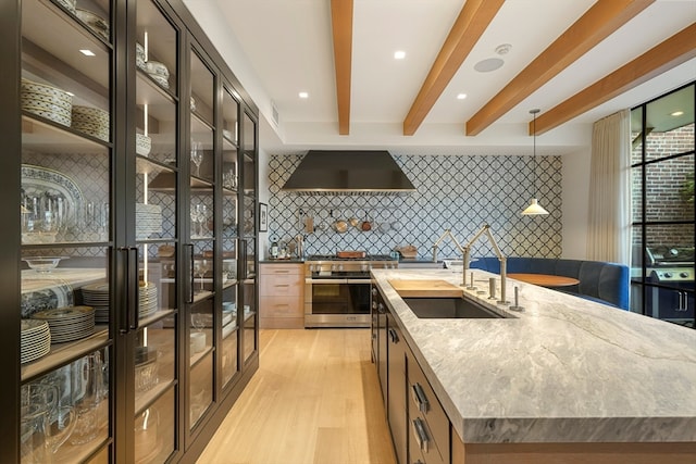 kitchen featuring decorative backsplash, an island with sink, wall chimney exhaust hood, high end range, and sink