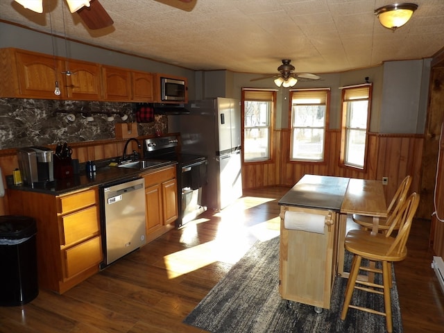 kitchen with ceiling fan, sink, dark wood-type flooring, stainless steel appliances, and wooden walls