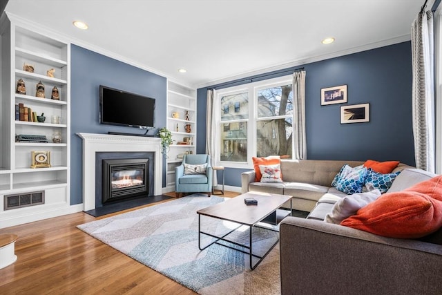 living area with built in shelves, wood finished floors, visible vents, ornamental molding, and a glass covered fireplace
