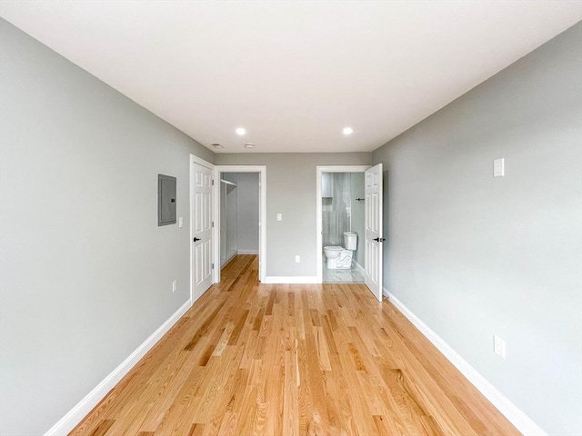 unfurnished bedroom with a spacious closet, connected bathroom, baseboards, light wood-type flooring, and electric panel