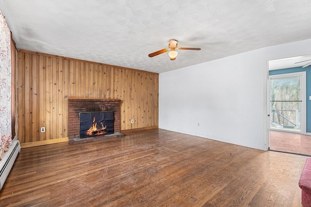 unfurnished living room featuring baseboard heating, a ceiling fan, a brick fireplace, wood walls, and wood finished floors