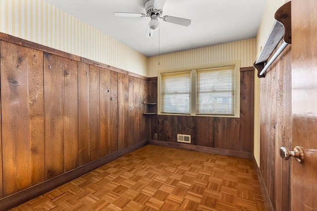 empty room with wooden walls, a ceiling fan, visible vents, baseboards, and wallpapered walls