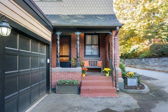 entrance to property featuring a garage, covered porch, brick siding, and roof with shingles