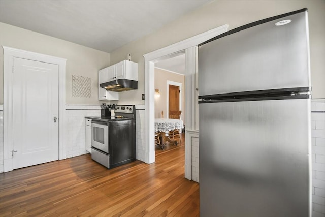 kitchen featuring a wainscoted wall, white cabinets, under cabinet range hood, appliances with stainless steel finishes, and dark wood-style floors