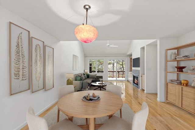 dining space with baseboards, light wood finished floors, ceiling fan, a lit fireplace, and vaulted ceiling