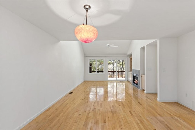 unfurnished living room featuring baseboards, lofted ceiling, light wood-style flooring, a warm lit fireplace, and a ceiling fan