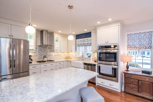 kitchen featuring appliances with stainless steel finishes, white cabinetry, a breakfast bar area, hanging light fixtures, and wall chimney range hood