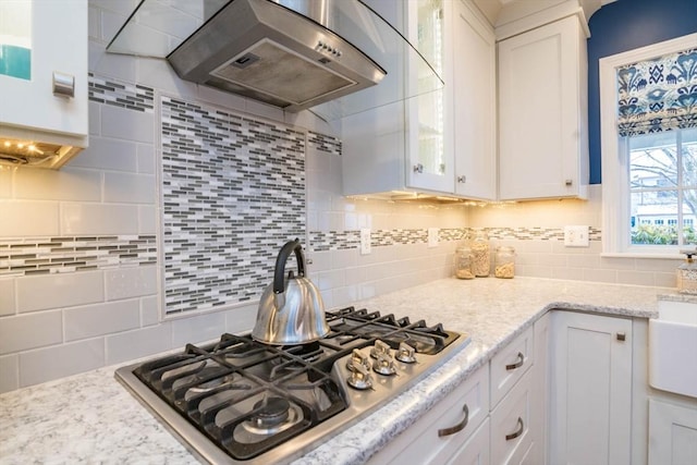 kitchen featuring wall chimney range hood, light stone countertops, stainless steel gas cooktop, white cabinets, and decorative backsplash