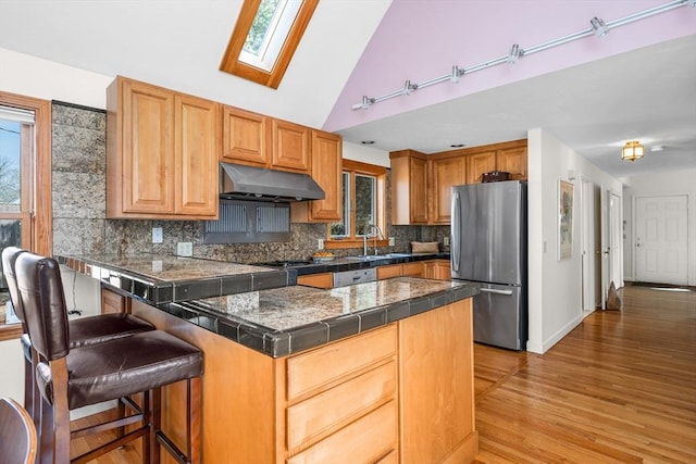 kitchen with lofted ceiling with skylight, under cabinet range hood, a sink, freestanding refrigerator, and a peninsula