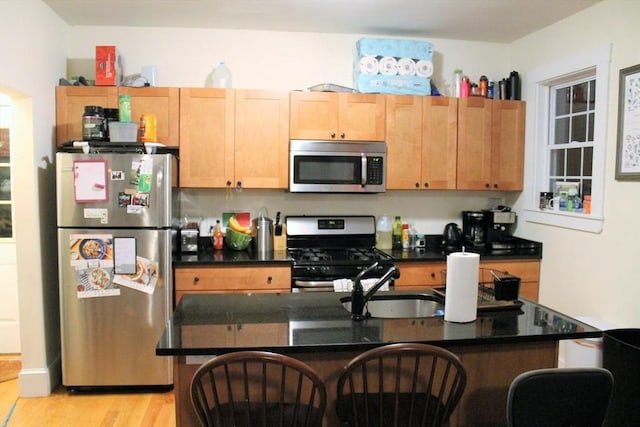 kitchen with light wood-style flooring, a kitchen island with sink, a sink, appliances with stainless steel finishes, and dark countertops