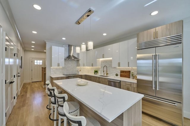 kitchen featuring light wood finished floors, appliances with stainless steel finishes, a kitchen breakfast bar, wall chimney range hood, and a sink