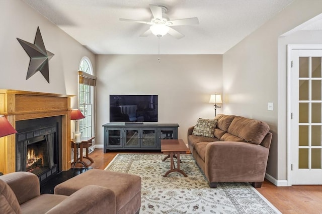 living area with baseboards, a ceiling fan, a warm lit fireplace, and wood finished floors