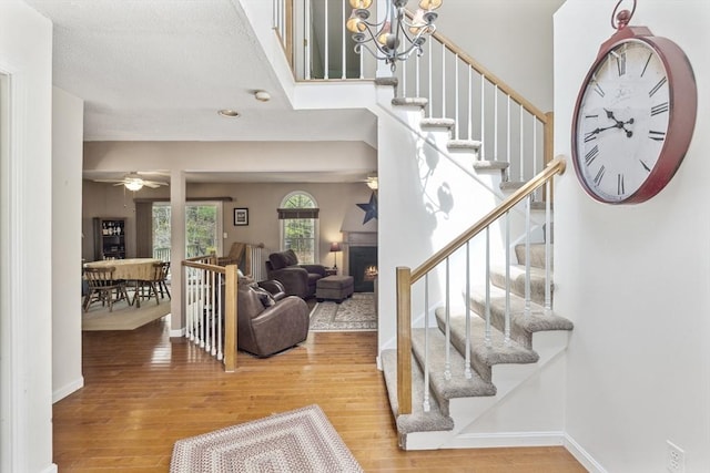foyer with stairway, a lit fireplace, wood finished floors, and ceiling fan with notable chandelier