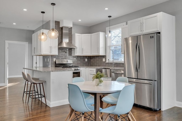 kitchen with appliances with stainless steel finishes, wall chimney exhaust hood, white cabinets, and hanging light fixtures