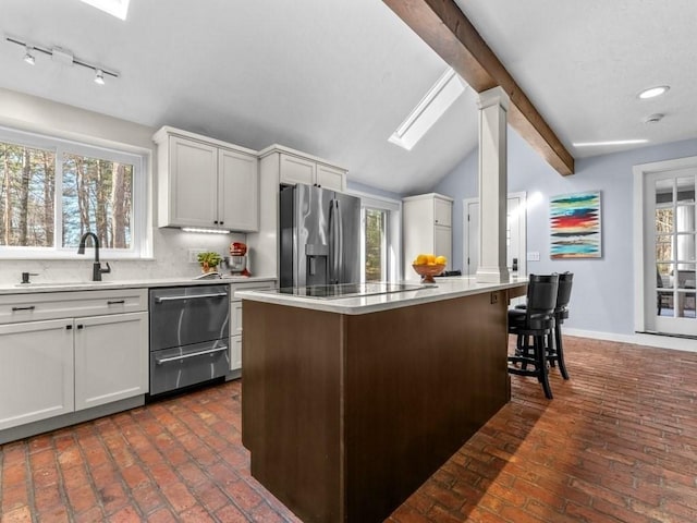 kitchen featuring backsplash, lofted ceiling with skylight, brick floor, stainless steel appliances, and a sink