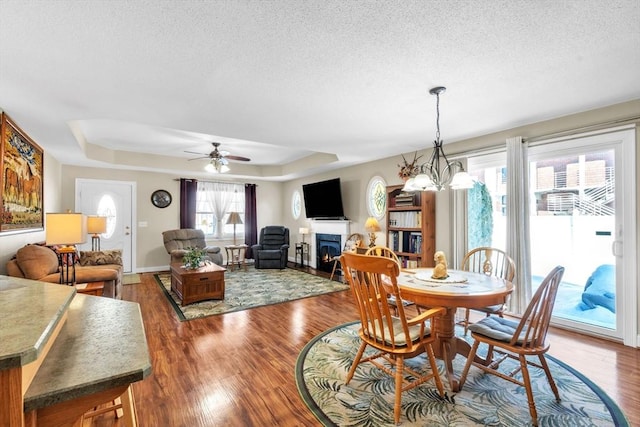 dining area with a warm lit fireplace, baseboards, wood finished floors, a tray ceiling, and a textured ceiling