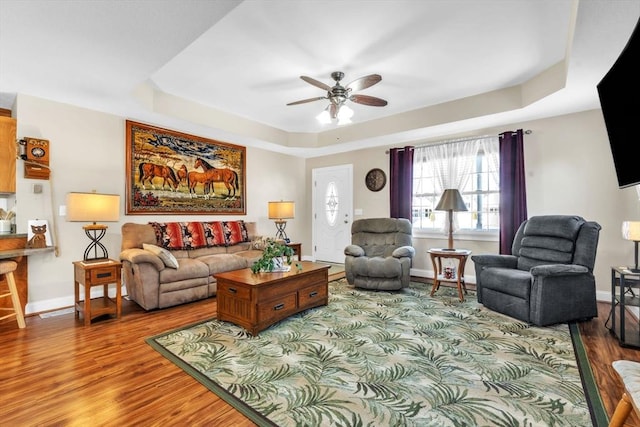 living area with a ceiling fan, baseboards, a tray ceiling, and wood finished floors