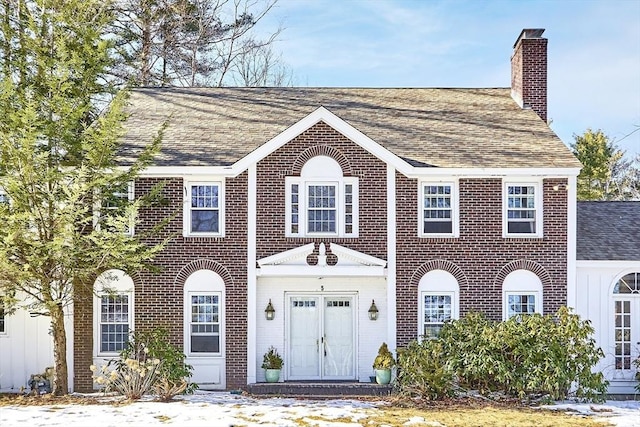 colonial house featuring roof with shingles, a chimney, and brick siding