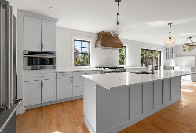 kitchen featuring premium range hood, an island with sink, hanging light fixtures, appliances with stainless steel finishes, and light hardwood / wood-style floors